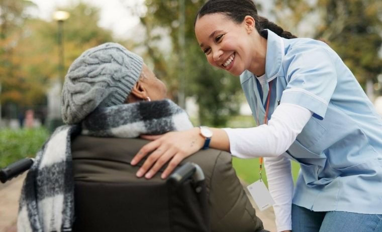 Smiling caregiver providing Personal Care in Michigan, offering compassionate support to an elderly woman in a wheelchair outdoors.